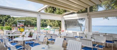 An outdoor patio with white chairs and tables, overlooking a beach and the sea.
