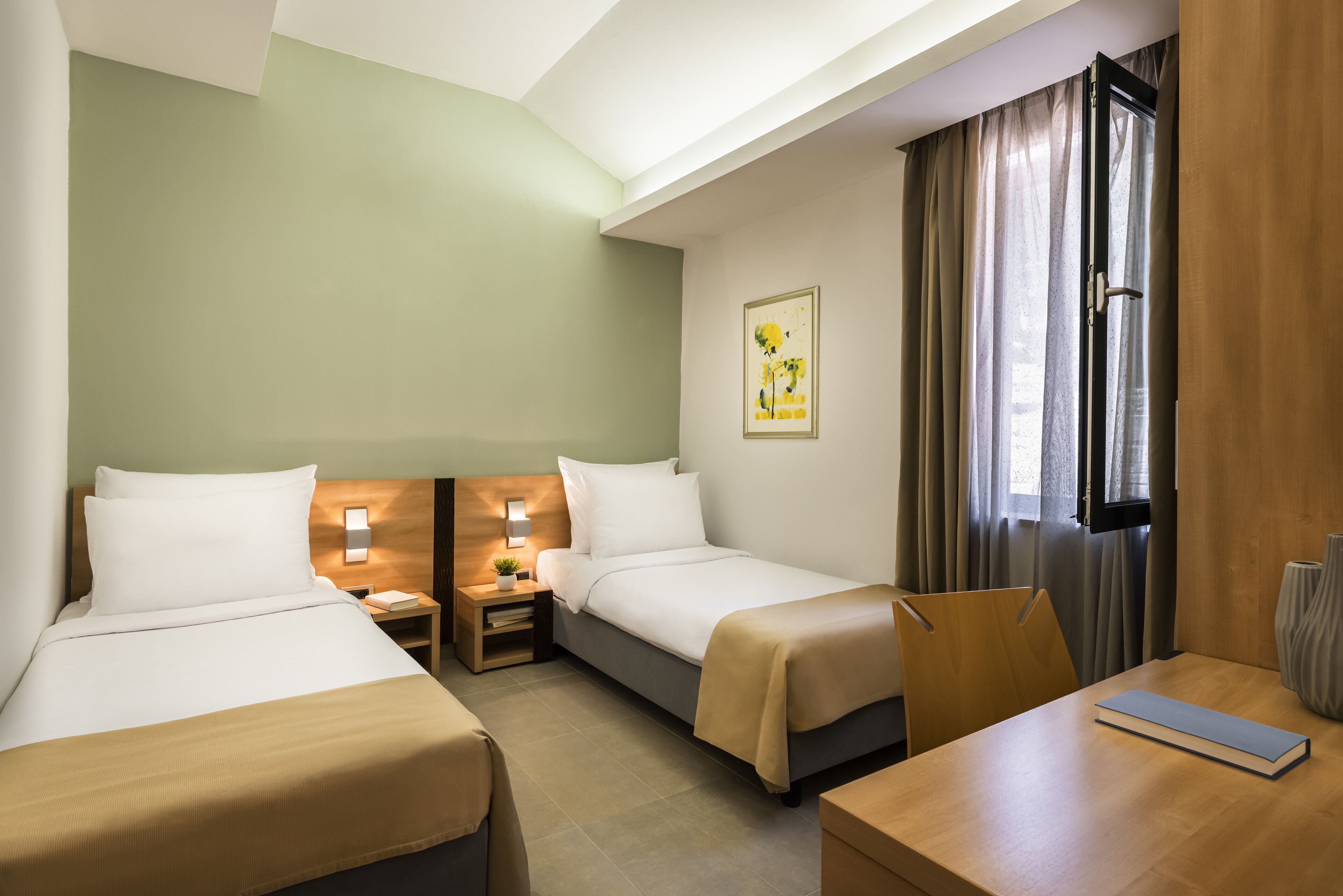 Ein Hotelzimmer mit zwei Betten, einem Holznachtkästchen mit Lampe, einem kleinen Tisch mit einem Buch und einem Fenster mit Vorhängen.