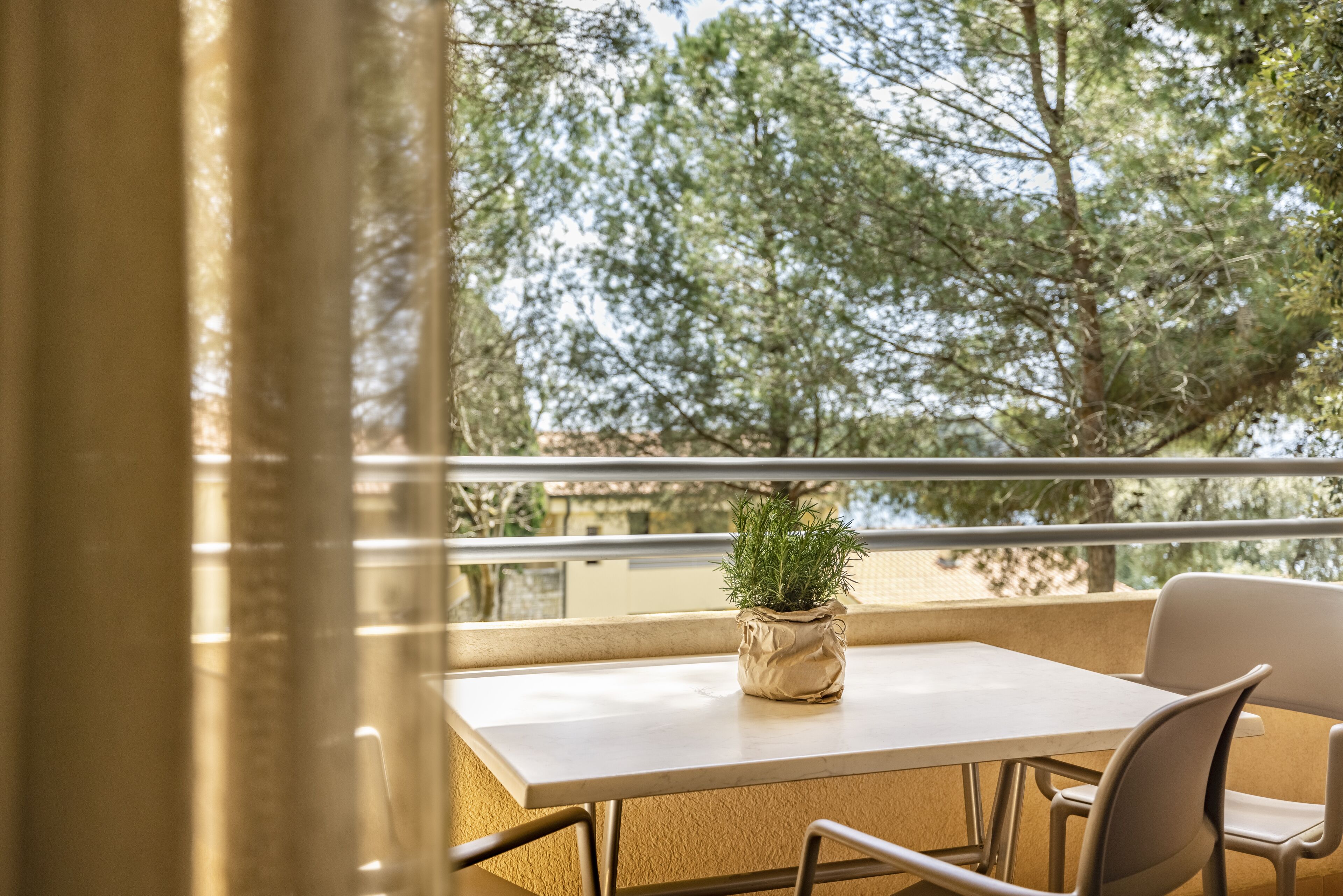 A balcony with a table and chairs, a potted plant, and a view of trees and buildings.