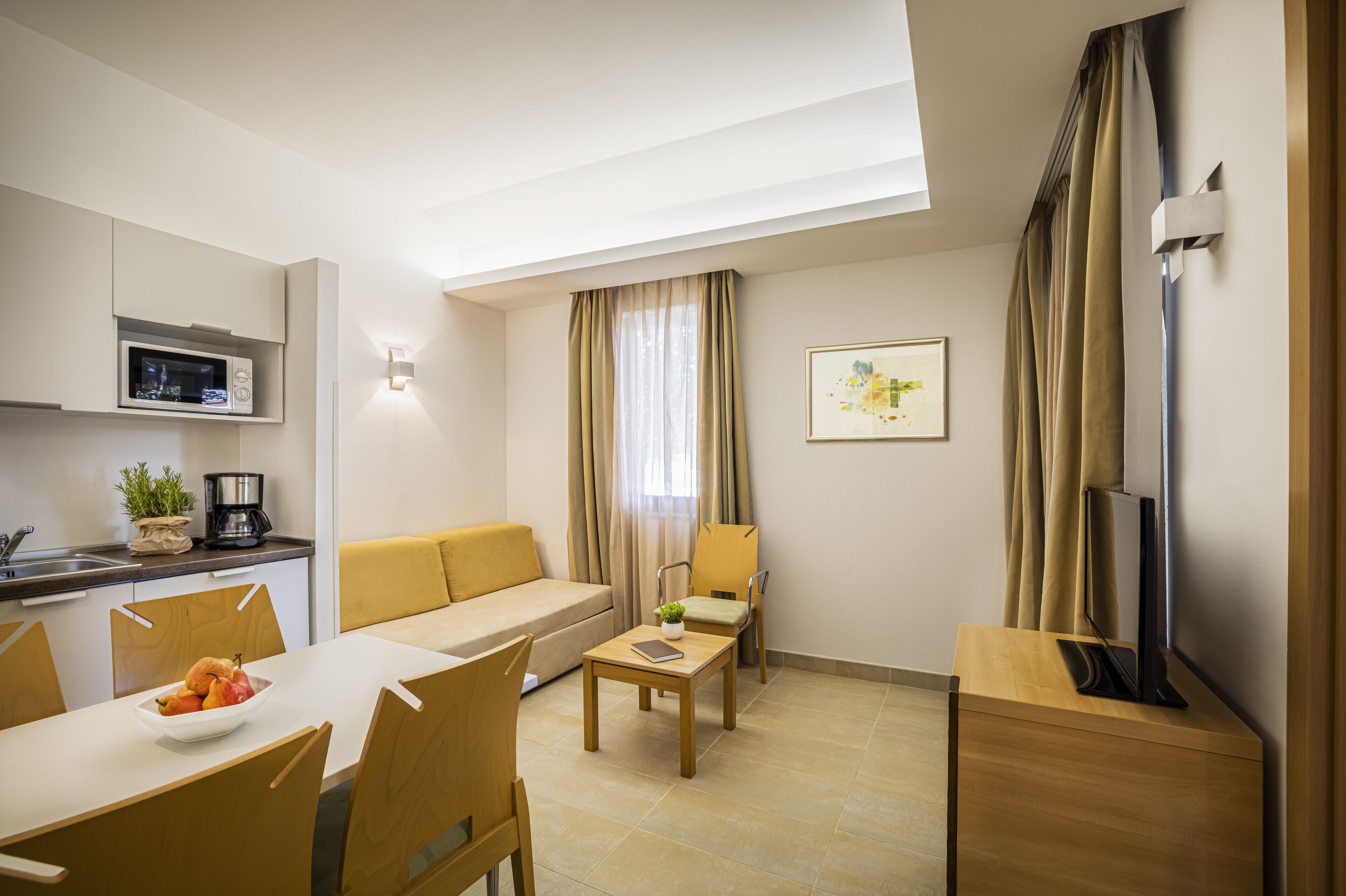 Habitación de hotel moderna con zona de comedor, sofá, televisión y kitchenette con microondas.