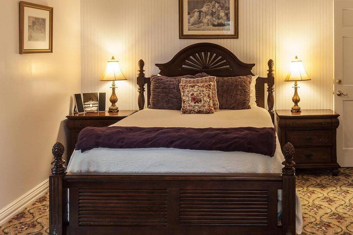 Ein traditionell eingerichtetes Schlafzimmer mit einem Himmelbett, zwei Nachttischen inklusive Lampen, einer floralen Tagesdecke und einem gerahmten Bild an der Wand.