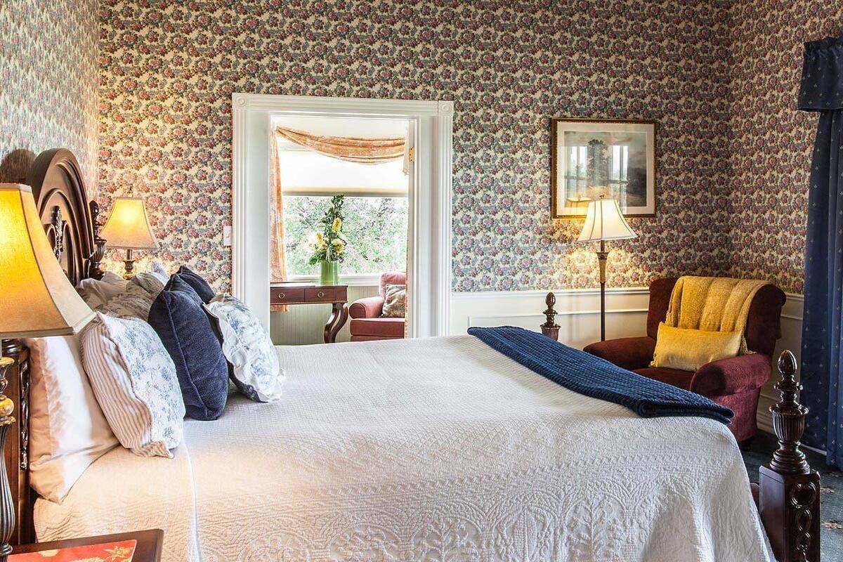 Ein Schlafzimmer mit einem großen Bett, zwei Nachttischlampen, einem Sessel mit gelber Decke, einem Fenster mit Vorhängen und blumigem Tapetenmuster.