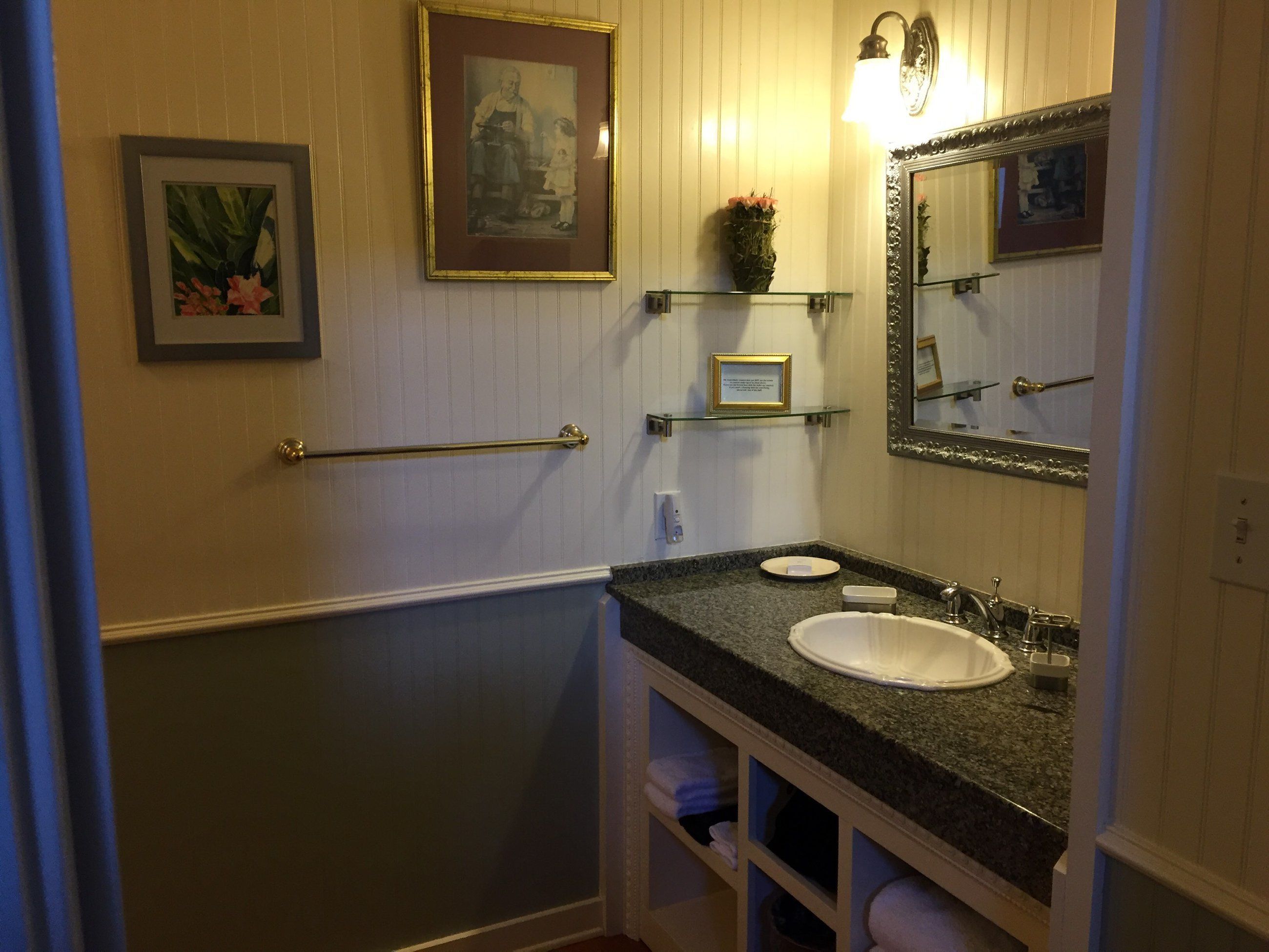 Ein Badezimmer mit Waschbecken, Spiegel, Handtuchhalter, gerahmten Bildern und einem kleinen Regal mit einer Vase.