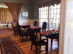 A dining area with wooden tables and chairs, a patterned rug, and large windows with curtains. - Rose Hill Retreat (Fredericksburg)
