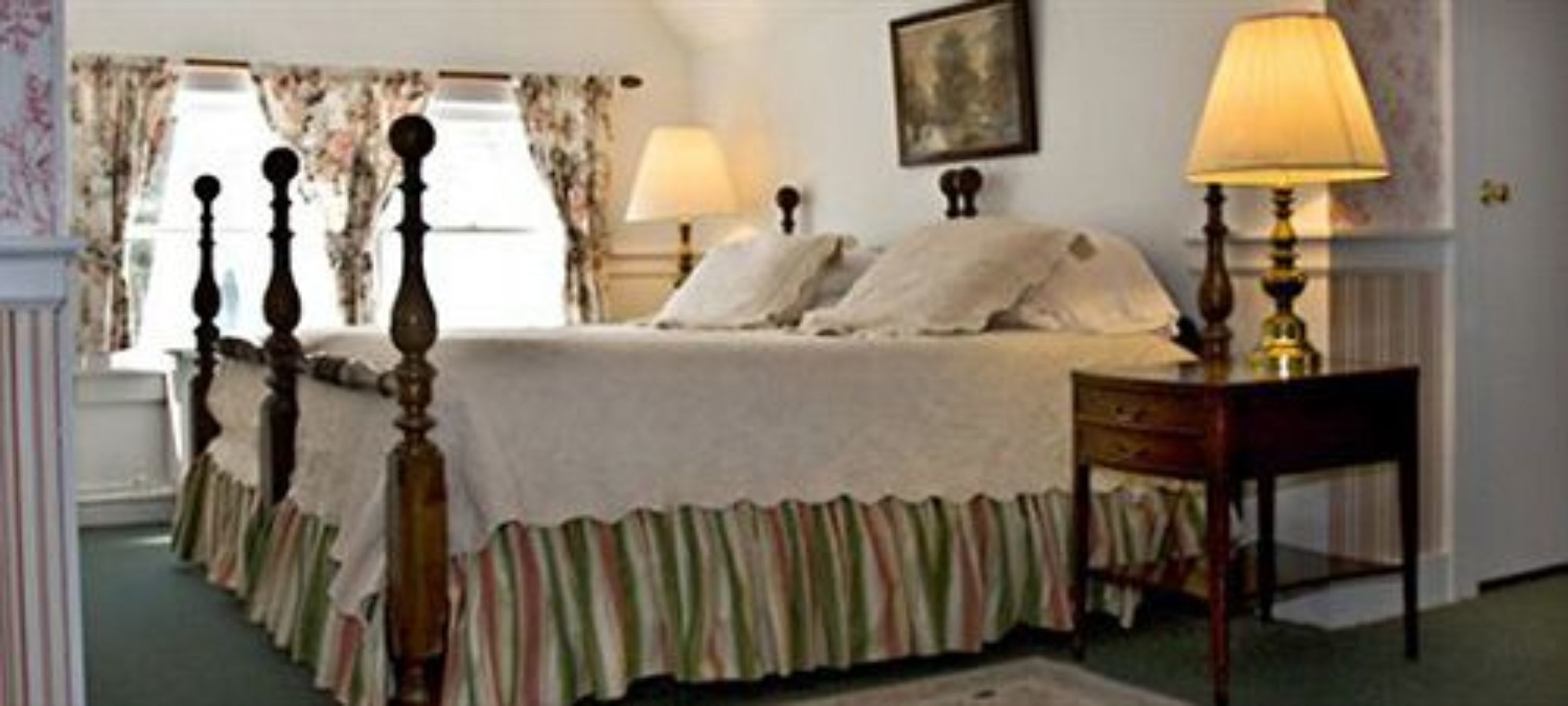 A bedroom with a bed, two bedside tables with lamps, a window with curtains, and a framed picture on the wall.