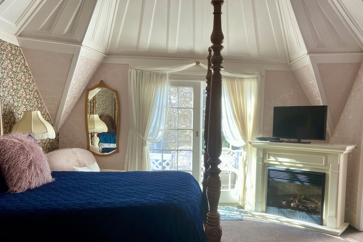 A bedroom with a four-poster bed, a television on a stand, a fireplace, and a balcony with curtains.