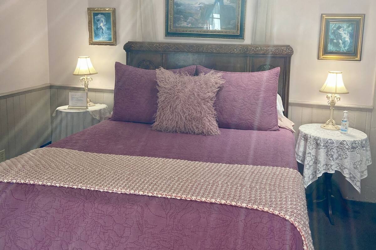 A neatly made bed with a purple bedspread and pillows, two bedside tables with lamps, and a framed painting above the bed.