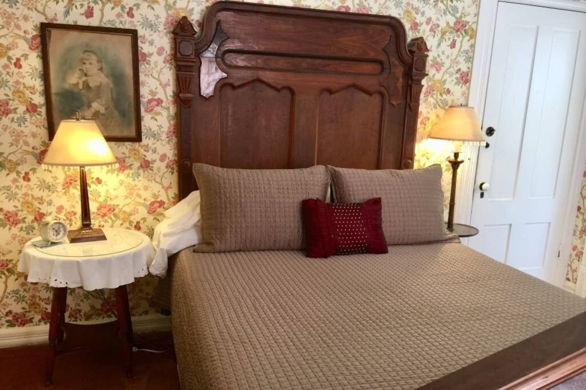 A bedroom with a wooden headboard, two bedside tables with lamps, floral wallpaper, and a bed with a quilted cover and two pillows.