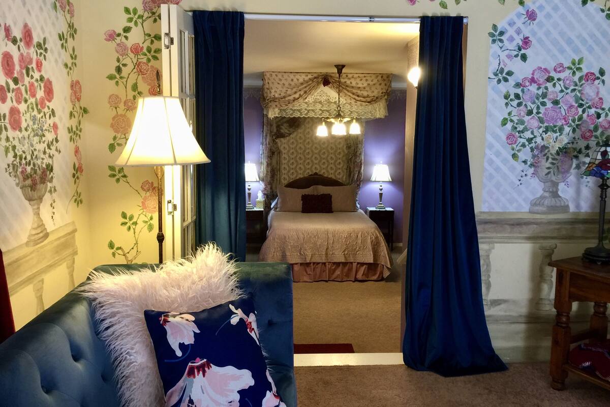 A hotel room with a bed, two bedside lamps, a blue armchair with a floral pillow, and floral wallpaper.