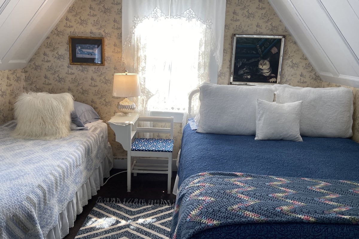 A cozy attic bedroom with two beds, a small table, and a window with lace curtains.