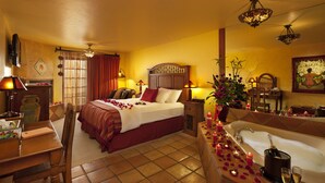 Una camera d'albergo con un letto, una zona salotto con un tavolo e sedie, una vasca da bagno con petali di rosa e uno specchio.
