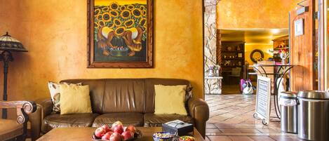 A living room with a brown leather sofa, a coffee table with fruits and bowls, a painting of sunflowers, and a kitchen area with a trash bin and a sign.