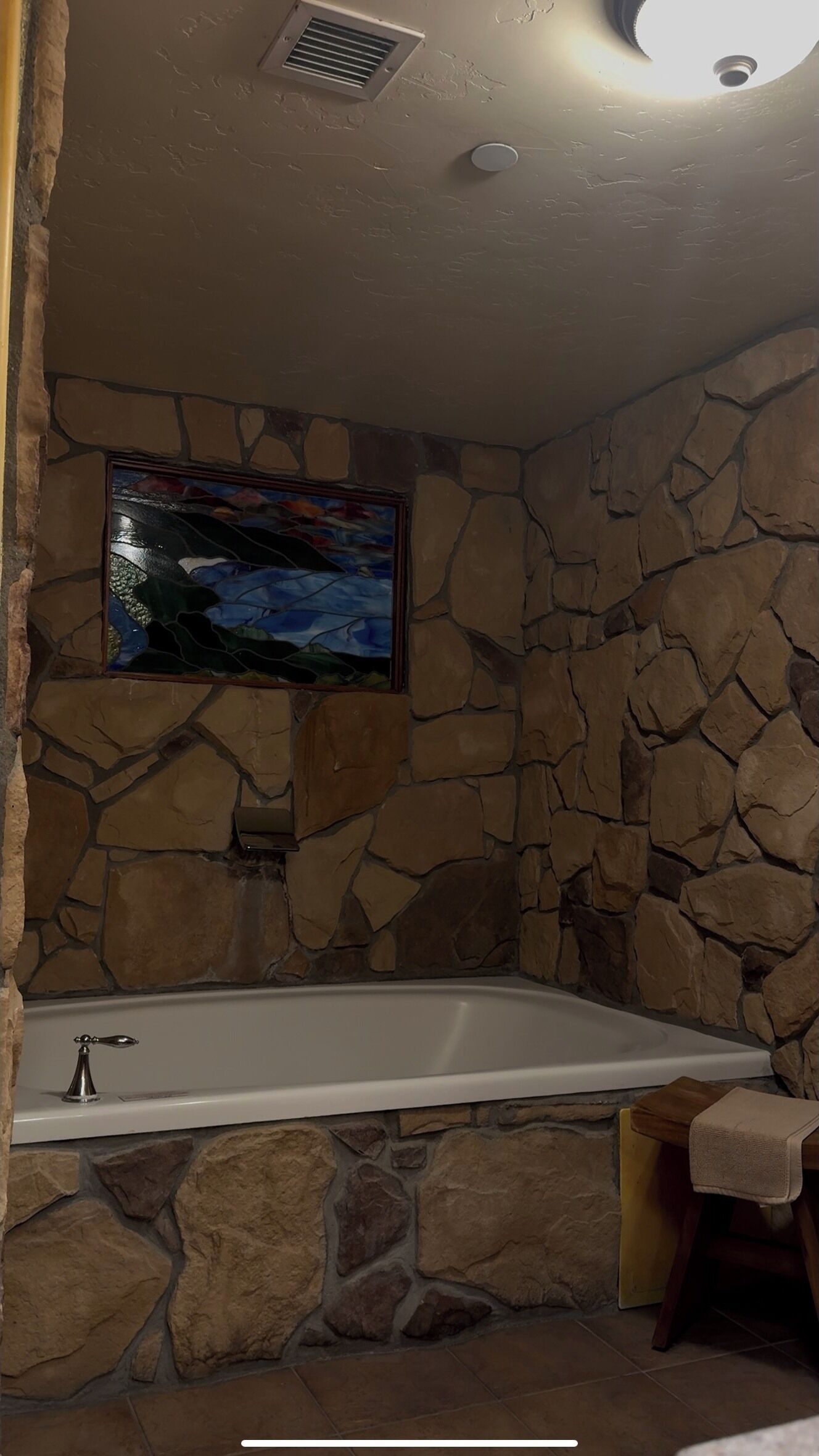 A stone-walled bathroom with a bathtub, a window with a stained glass design, and a towel rack.