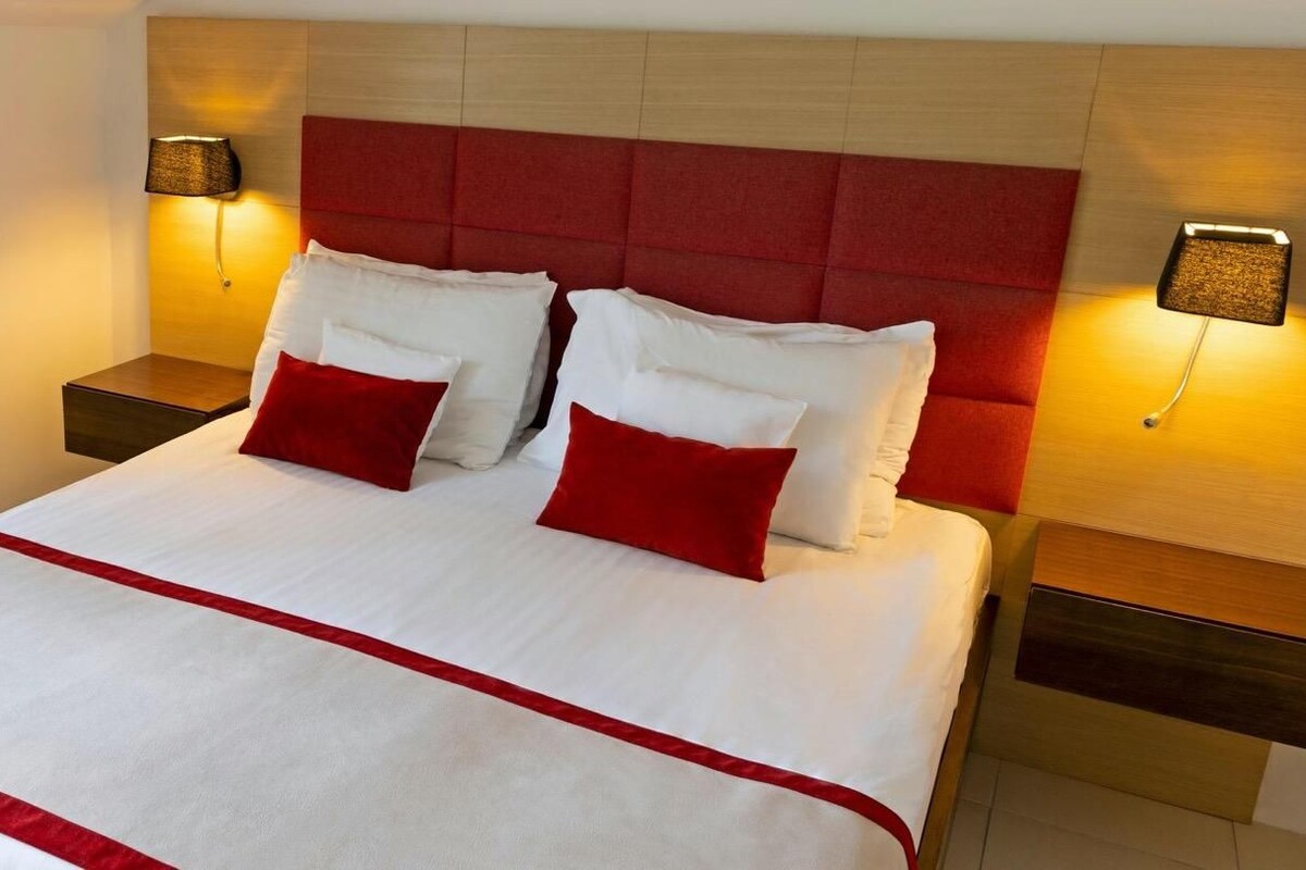 A hotel room with a large bed, red headboard, white bedding, and two bedside lamps.