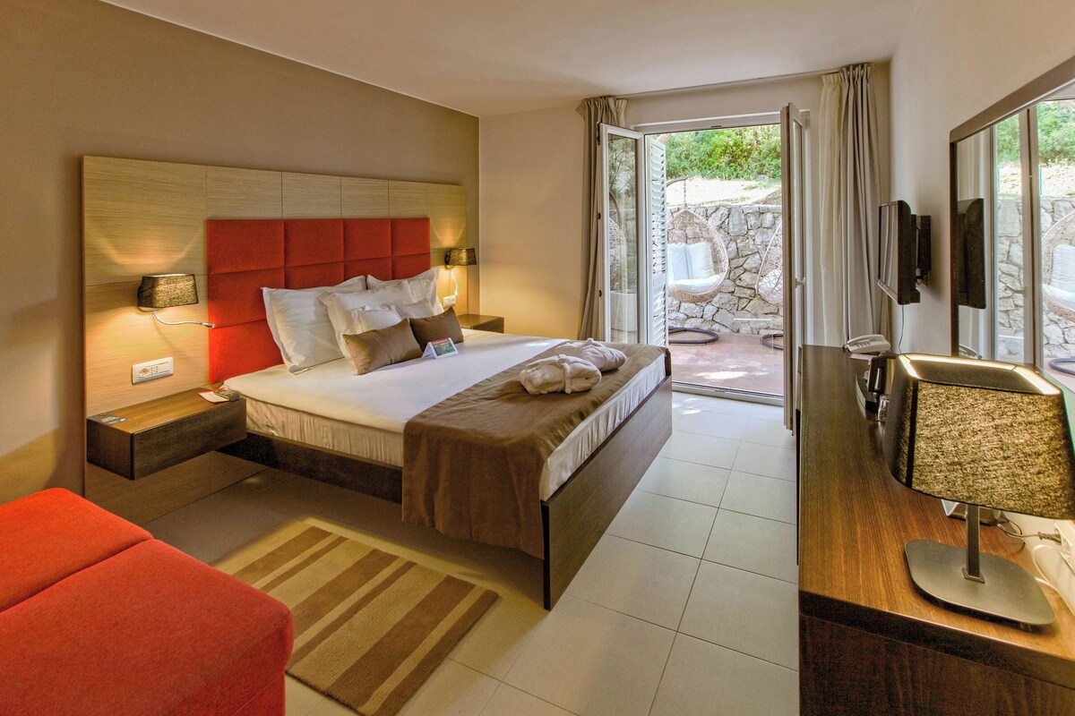 A modern hotel room with a large bed, a wooden headboard, a bedside table with a lamp, a red armchair, and a view of the outdoor area through a glass door.