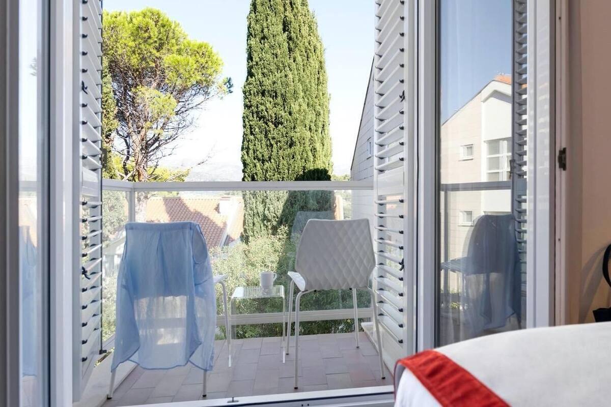 Un balcon donnant sur des arbres et des immeubles, une chaise blanche, une table avec une tasse, et un lit agrémenté d’une touche de rouge.