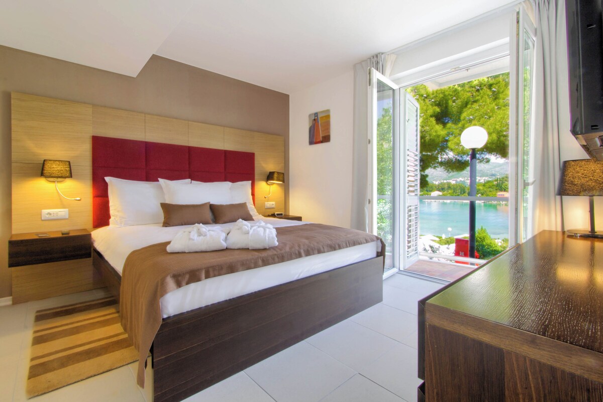 A modern hotel room with a large bed, a wooden headboard, a nightstand with a lamp, and a view of the sea through the window.