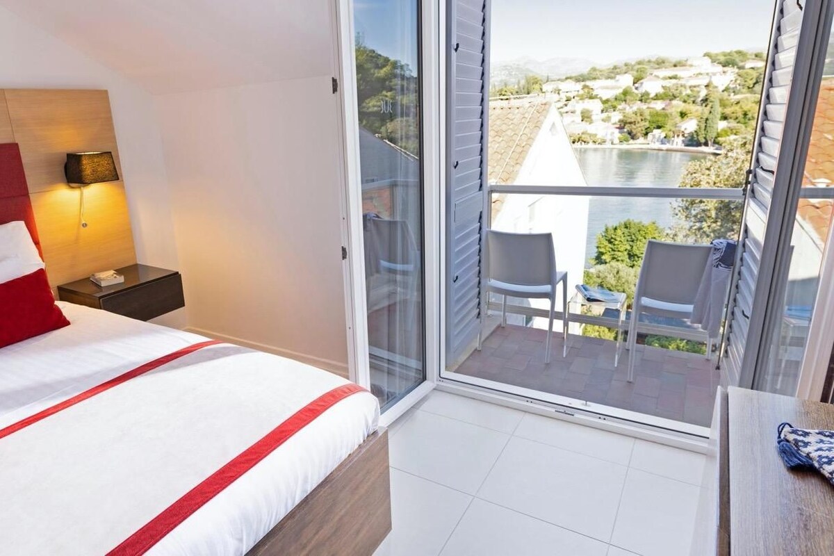 A hotel room with a bed, a nightstand, a lamp, a balcony with chairs, and a view of the landscape.