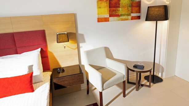 A hotel room with a bed, a chair, a nightstand, a lamp, and a wall art piece. - TUI Blue Kalamota Island (Dubrovnik)
