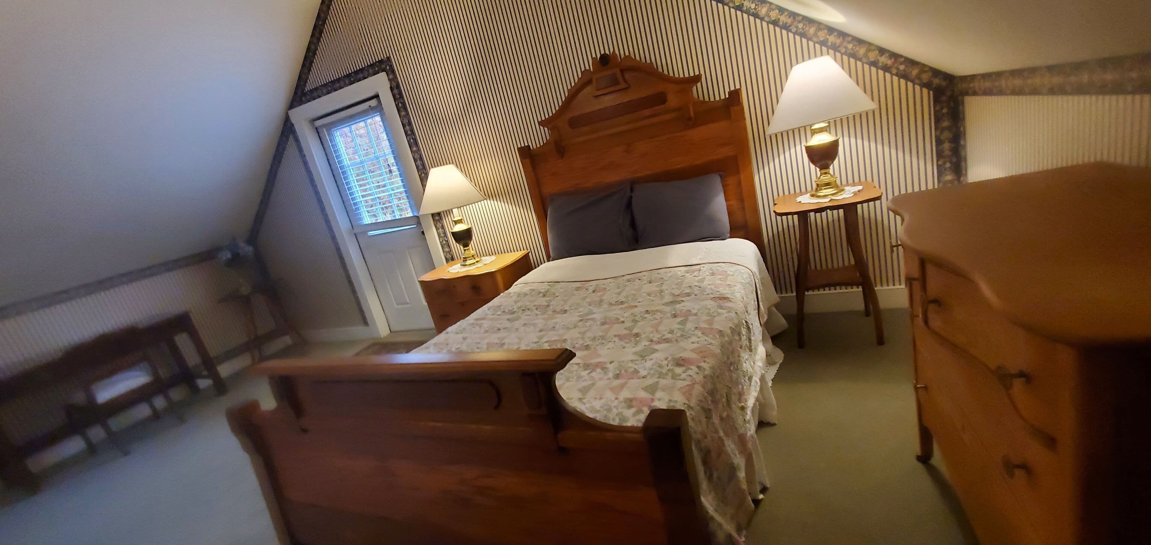 A bedroom with a sloped ceiling, a wooden bed, a nightstand with a lamp, a dresser, and a window with blinds.