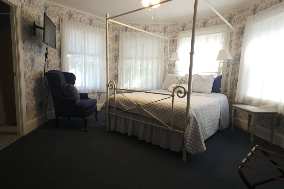 A four-poster bed with a canopy, a chair, a bedside table, and a window with curtains.