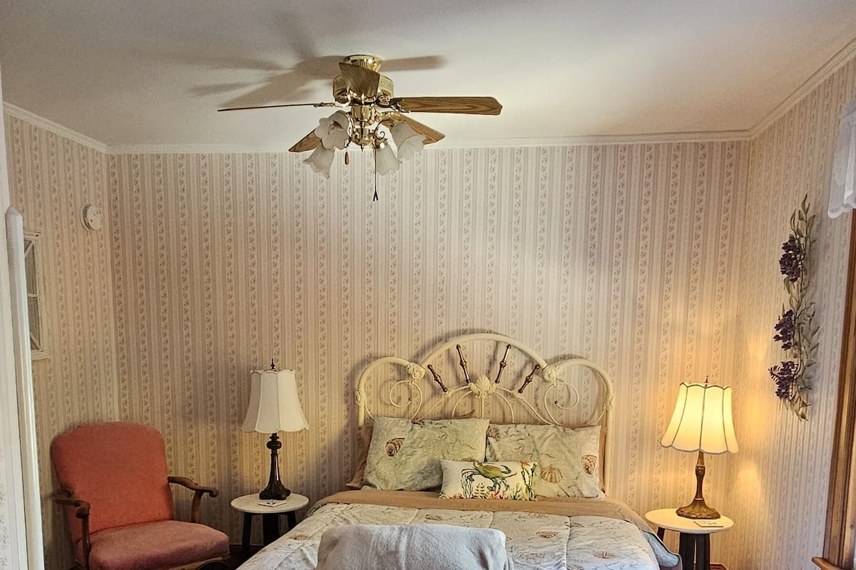 A hotel room with a large bed, two bedside tables with lamps, a ceiling fan, a red armchair, and a dresser.
