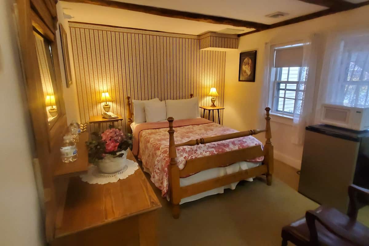 A bedroom with a four-poster bed, bedside lamps, a wooden nightstand with a vase of flowers, a dresser, and a window with curtains.
