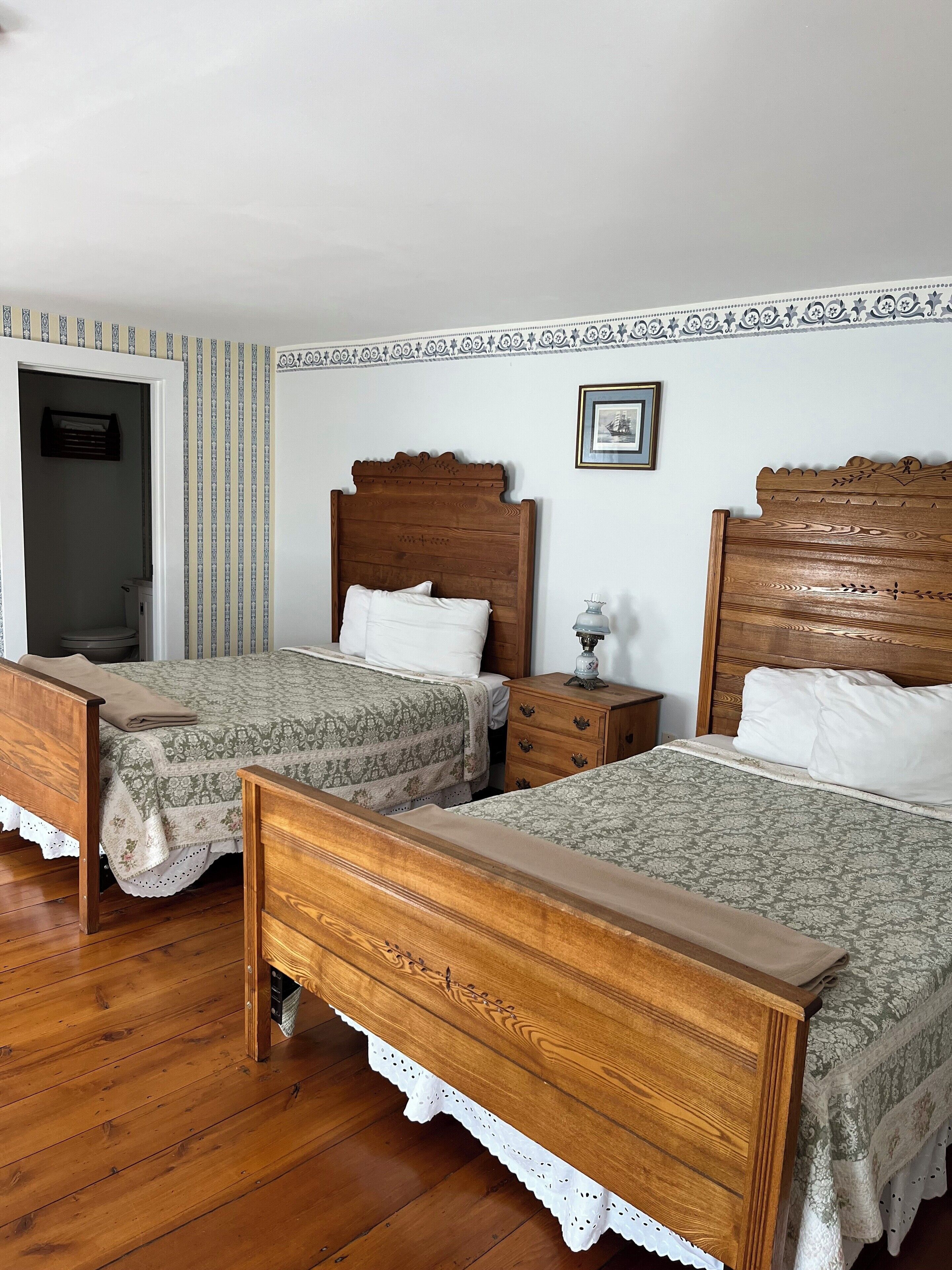 A room with two wooden beds, a wooden nightstand, a small table with a lamp, and a framed picture on the wall.