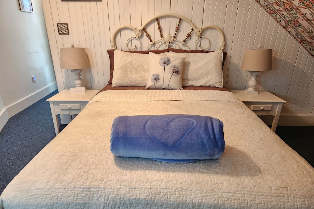 A neatly made bed with a blue heart-shaped pillow, white linens, and two bedside lamps on matching nightstands.