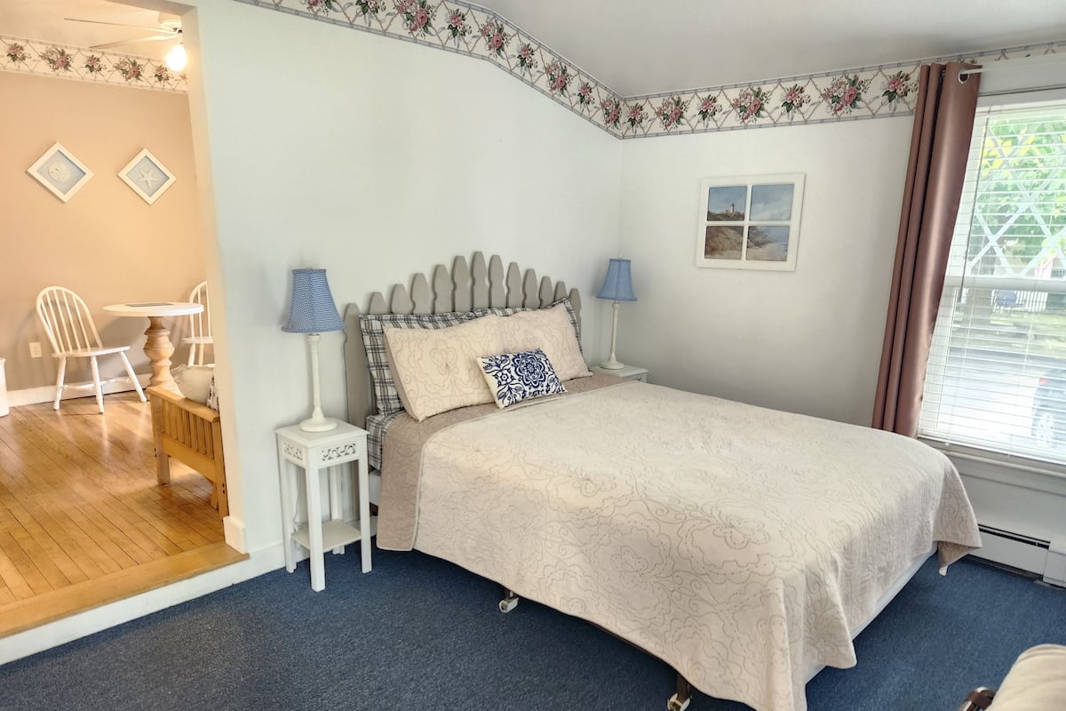 A bedroom with a bed, two bedside tables with lamps, a chair, a small table, and a window with curtains.