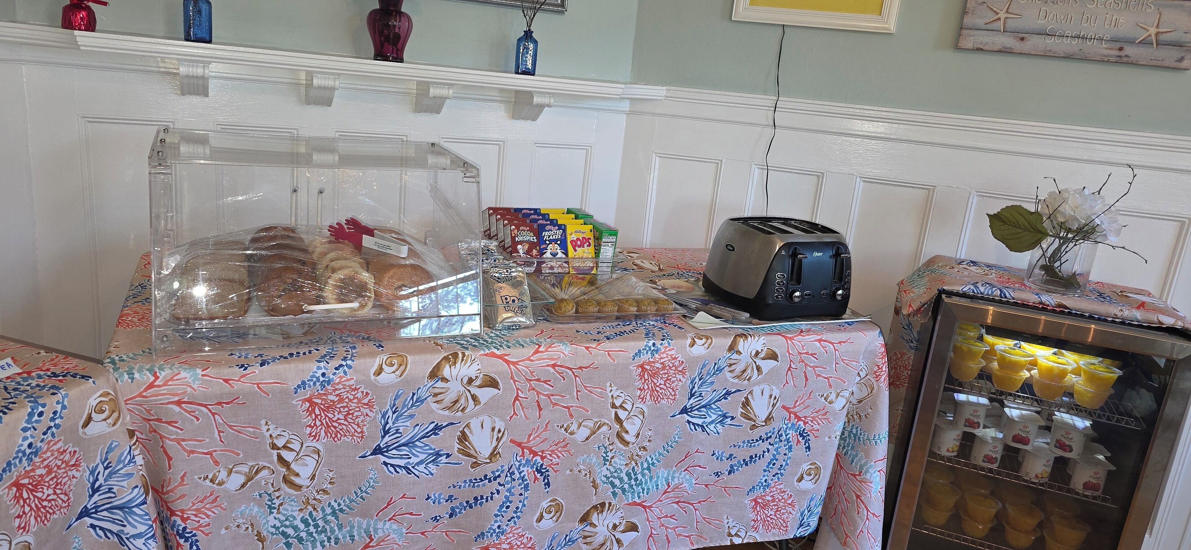 A breakfast setup with a covered bread display, cereal boxes, a toaster, and a refrigerator with beverages.