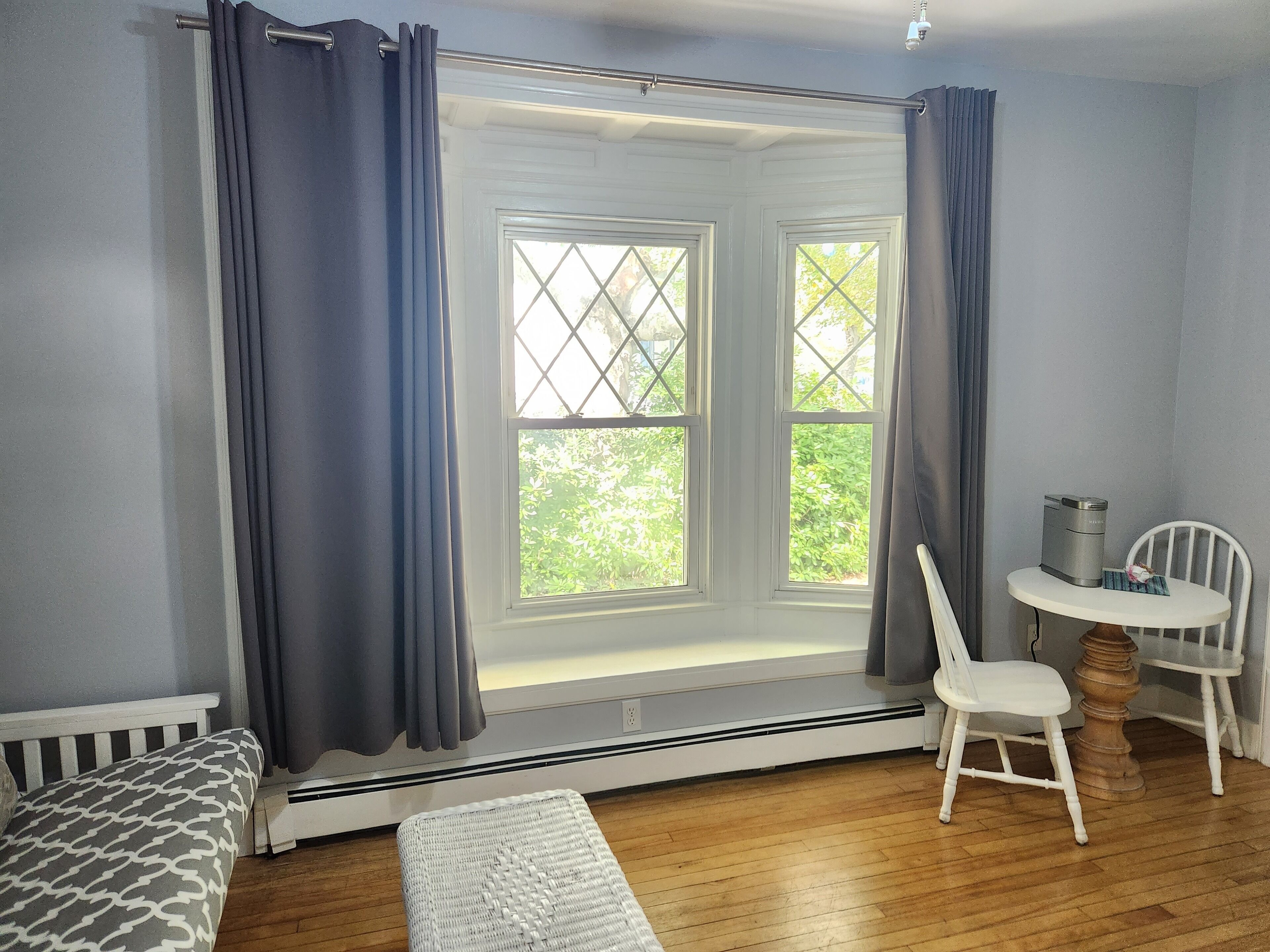 A room with a bay window, a white chair, a small table with a kettle, and a white bench.