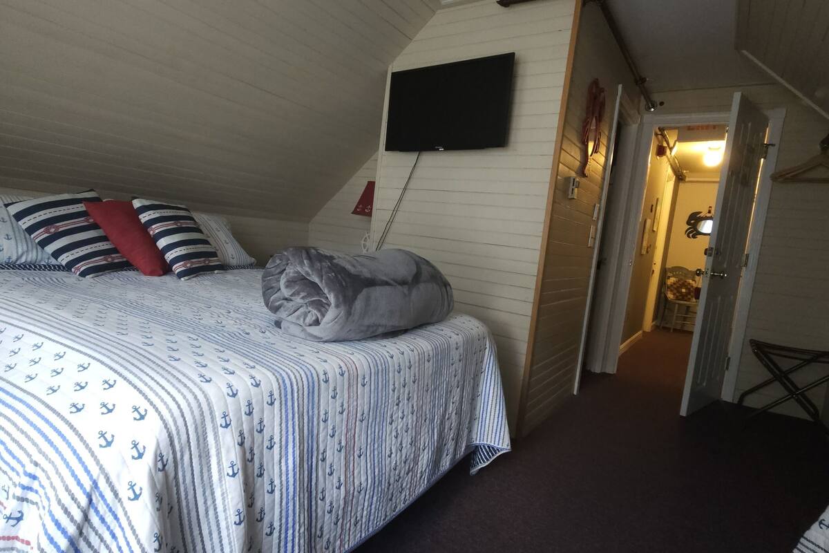 A bedroom with a bed, pillows, a blanket, a TV, and a door leading to another room.
