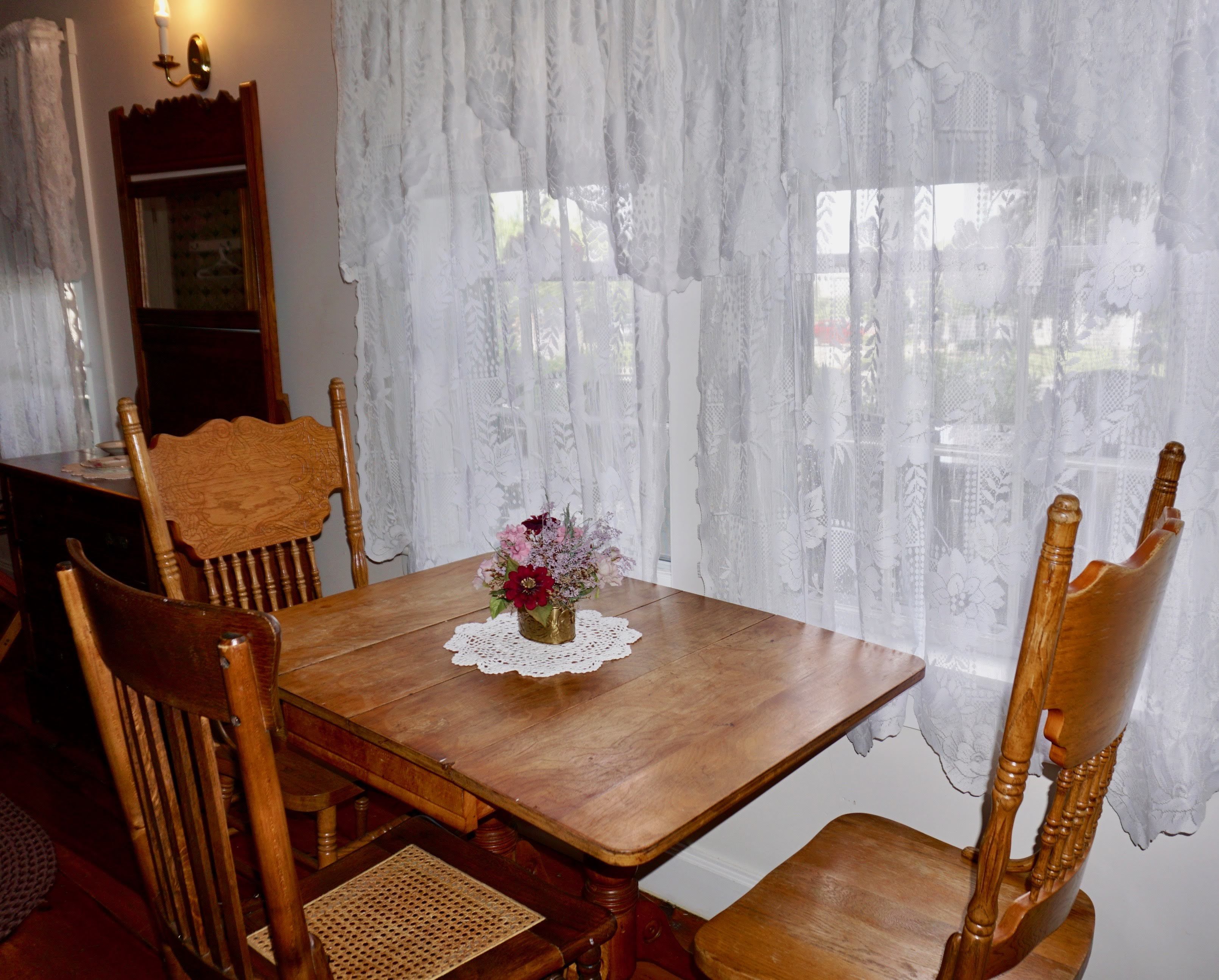Una mesa de comedor de madera con un posavasos de encaje, rodeada de sillas de madera con cojines de asiento tejidos. Un jarrón con flores está colocado sobre la mesa.