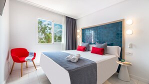Una habitación de hotel moderna con una cama grande, una silla roja, una mesita con un jarrón y vistas a los árboles a través de la ventana.