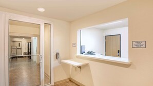 A hotel corridor with a glass door leading to a room, a check-in/check-out sign, and a small window with a view of the room. - WoodSpring Suites Jacksonville Beach Blvd (Jacksonville)
