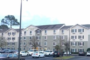A multi-story hotel with a sign reading 'Woodspring Suites.' There are several cars parked in the lot, and the building has a modern architectural style with a flat roof and multiple windows.