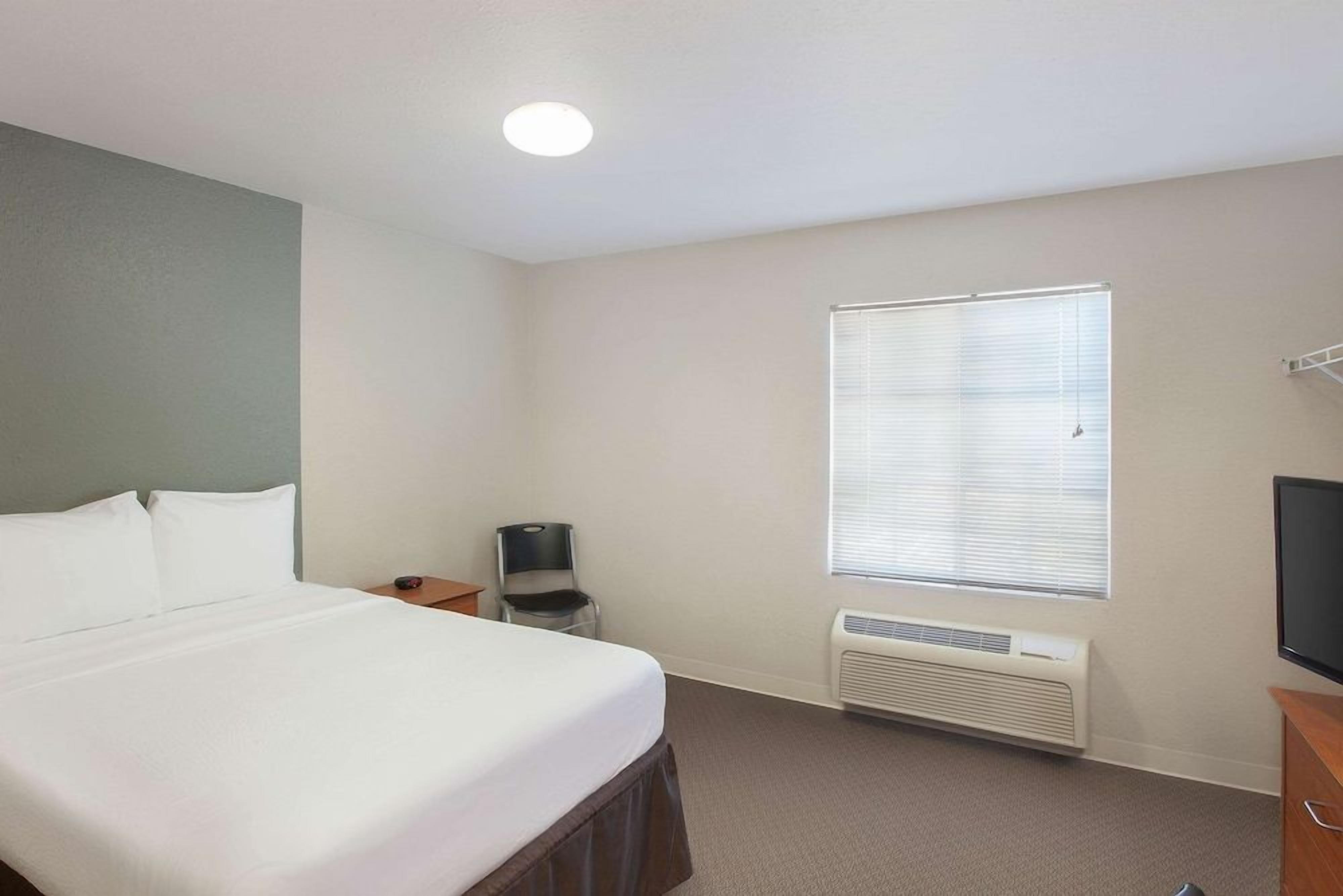 Una habitación de hotel con cama, una silla, un televisor, una ventana con persianas y un calentador de pared.