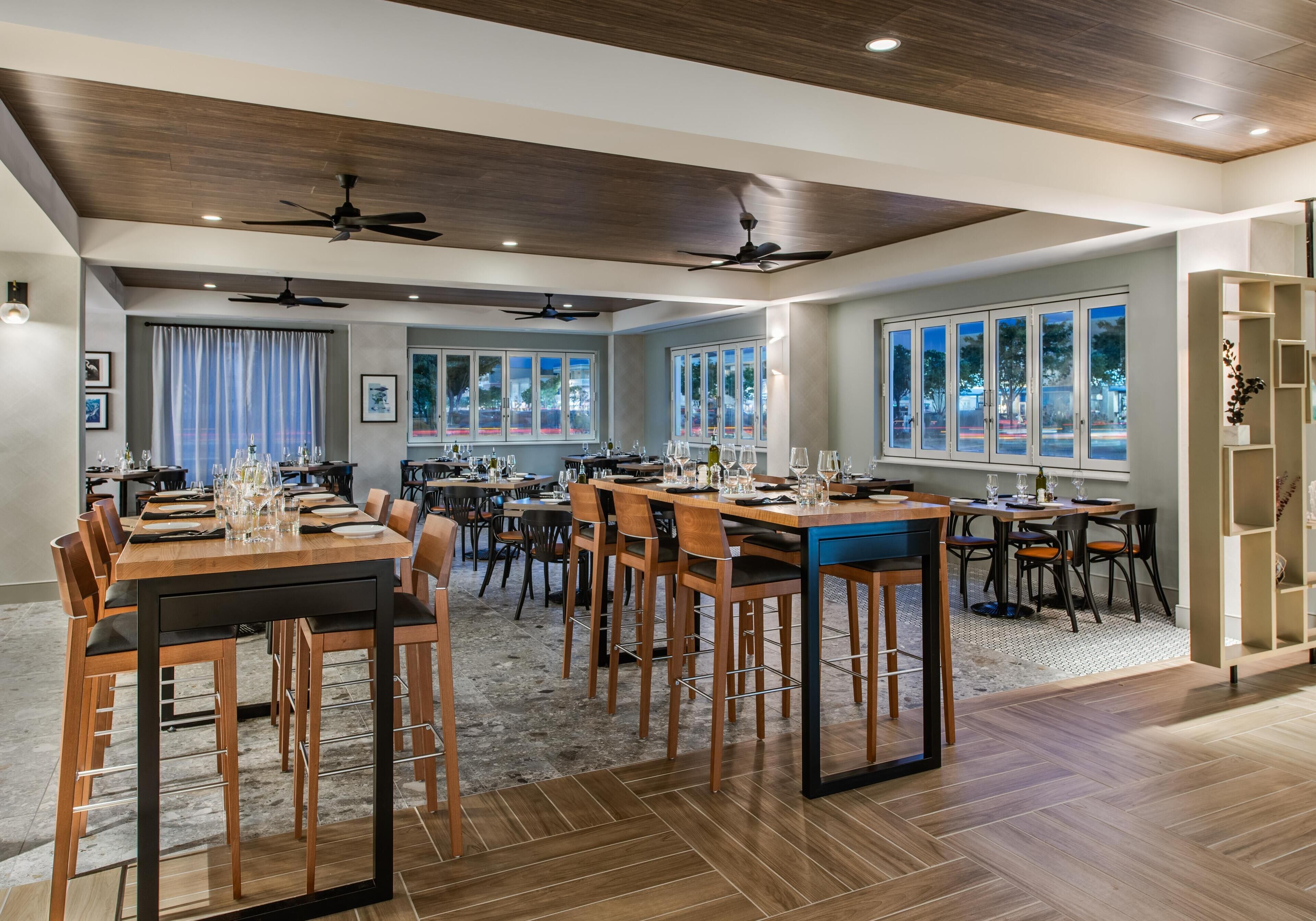 Un'ampia sala da pranzo con diversi tavoli e sedie, ventilatori a soffitto e grandi finestre che lasciano entrare la luce naturale.