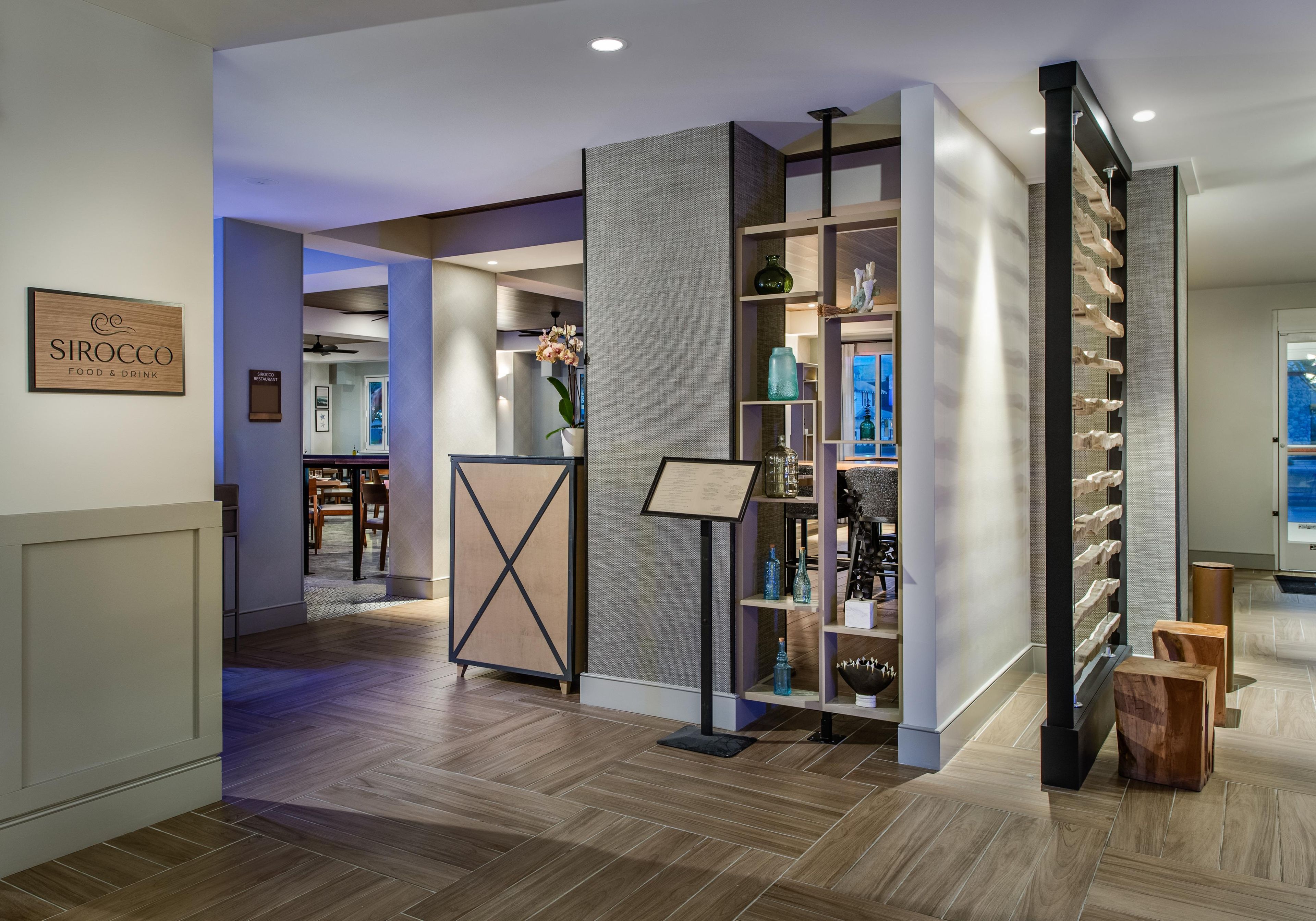 A modern interior with a wooden floor, a reception desk, and a wall-mounted sign for Sirocco Restaurant.