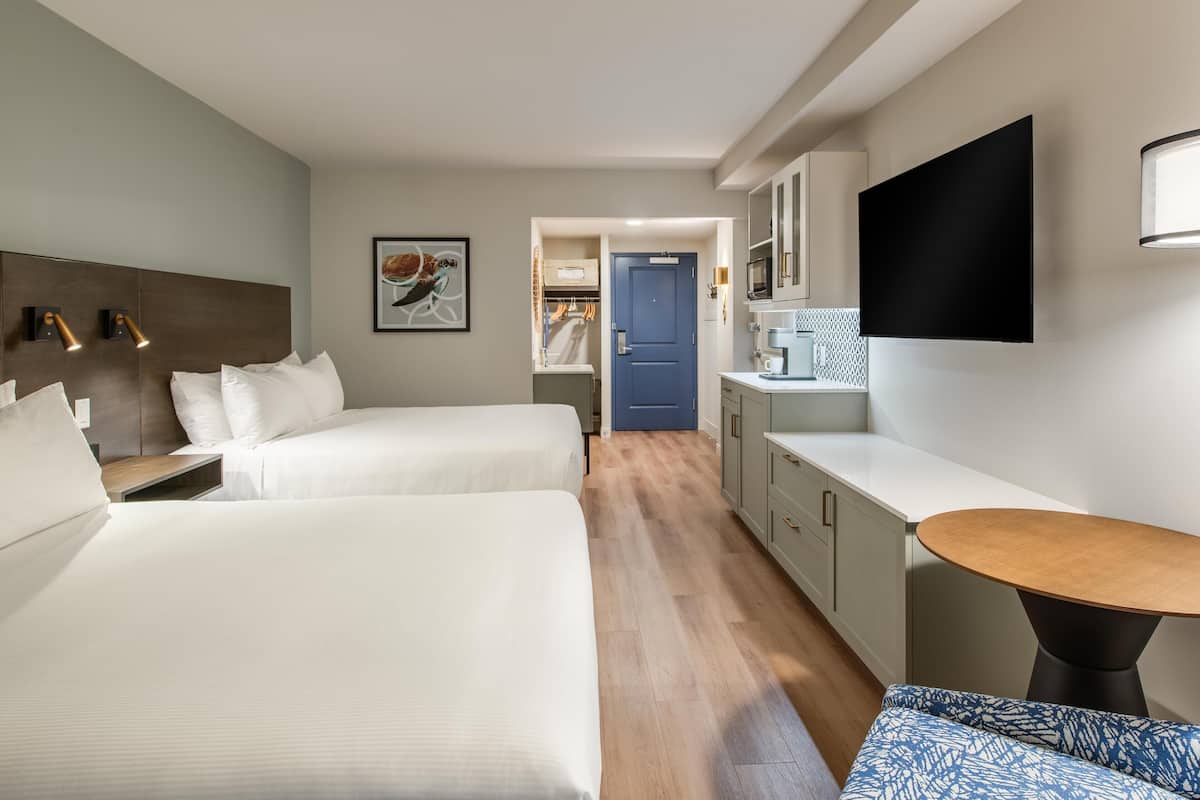 A hotel room with two beds, a small kitchenette, a TV mounted on the wall, and a wooden floor.