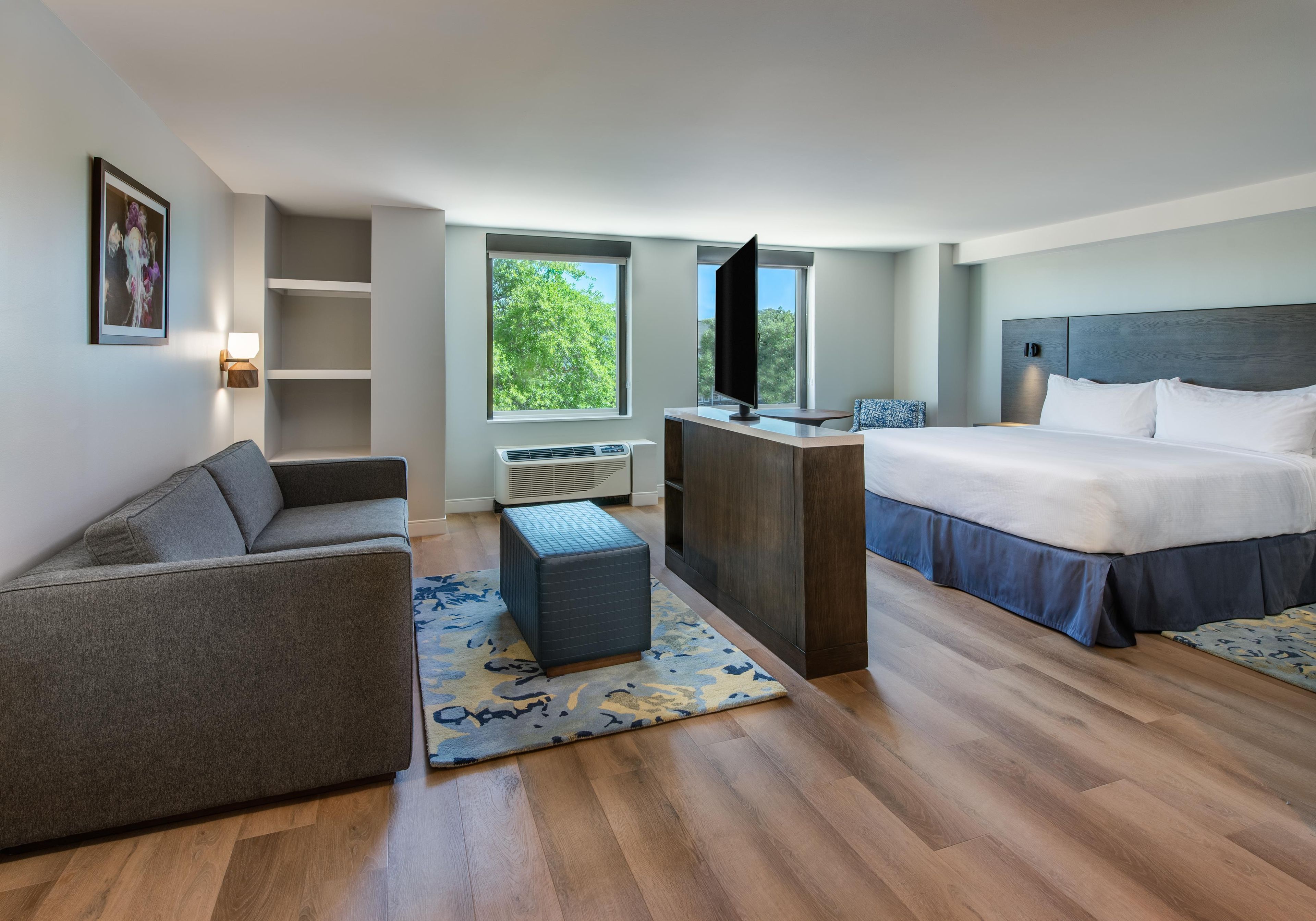A hotel room with a bed, a sofa, a TV, a desk, and a window with a view of trees.