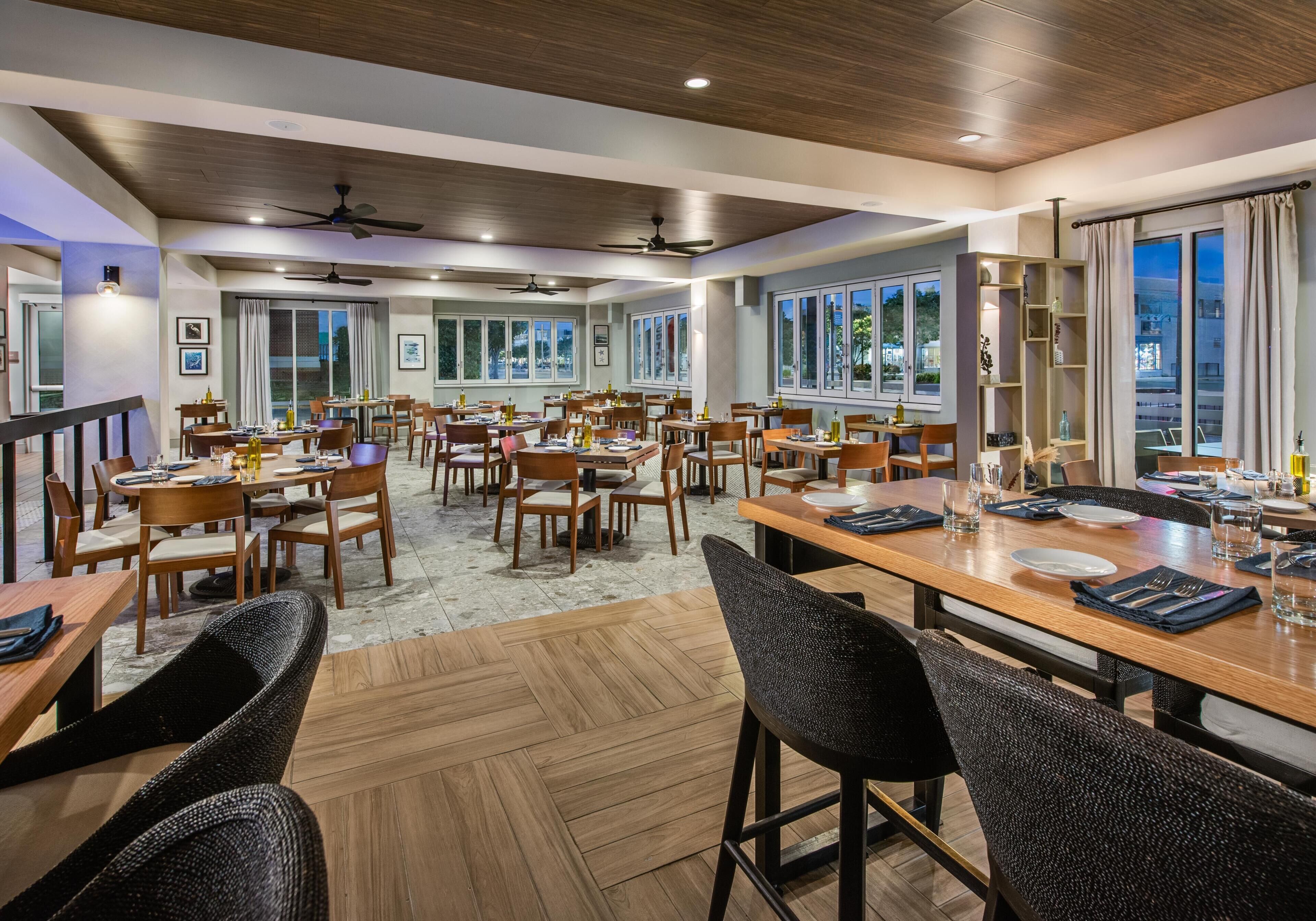 A spacious dining area with multiple tables and chairs, large windows, and ceiling fans.