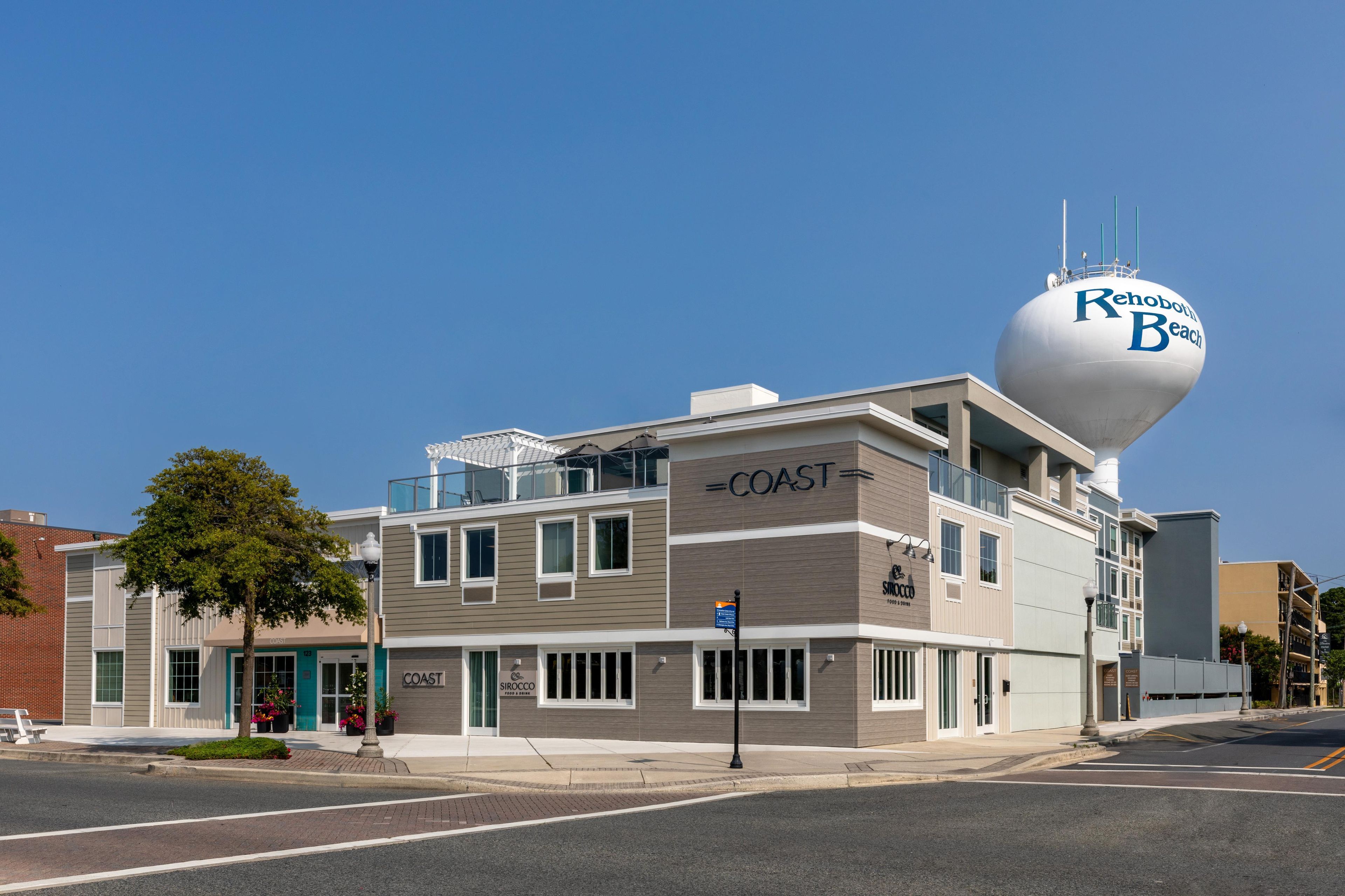 Un immeuble à plusieurs étages, surmonté d’un château d’eau bien visible portant l’inscription « Rehoboth Beach », et sur lequel on peut également voir une enseigne pour le restaurant « COAST ».