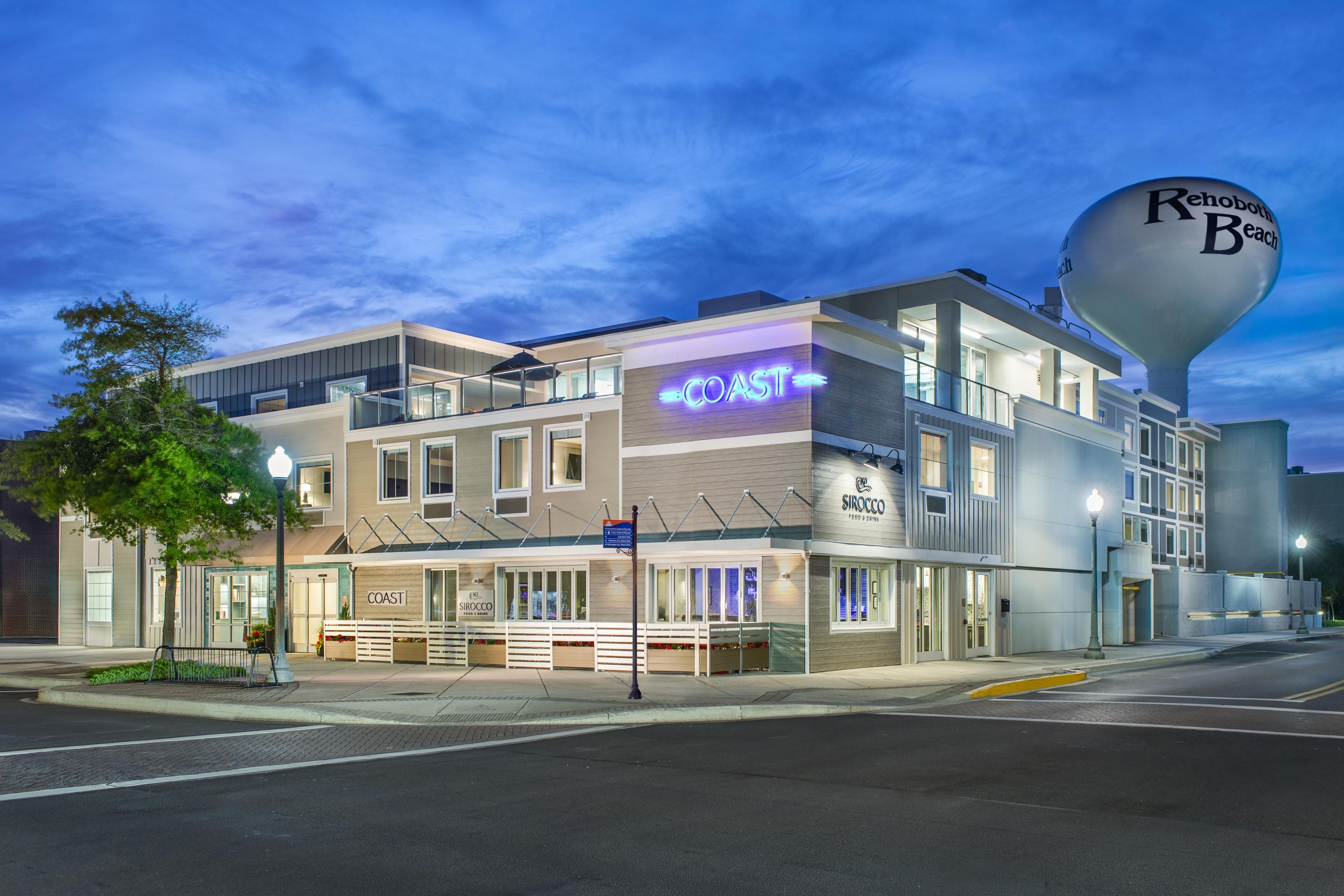 Un hotel de varios pisos con un cartel llamativo que dice 'COAST', una torre de agua con la leyenda 'Rehoboth Beach' y un letrero que indica 'SIROCCO'.