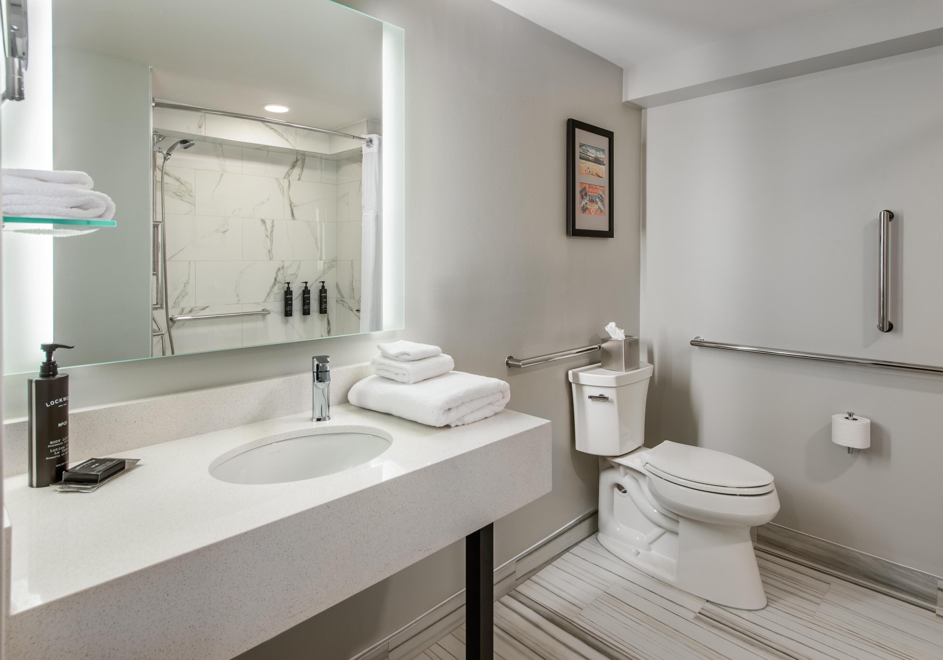 A modern bathroom with a white sink, mirror, and toilet. There is a towel rack with folded towels, a glass shower enclosure, and a framed picture on the wall.