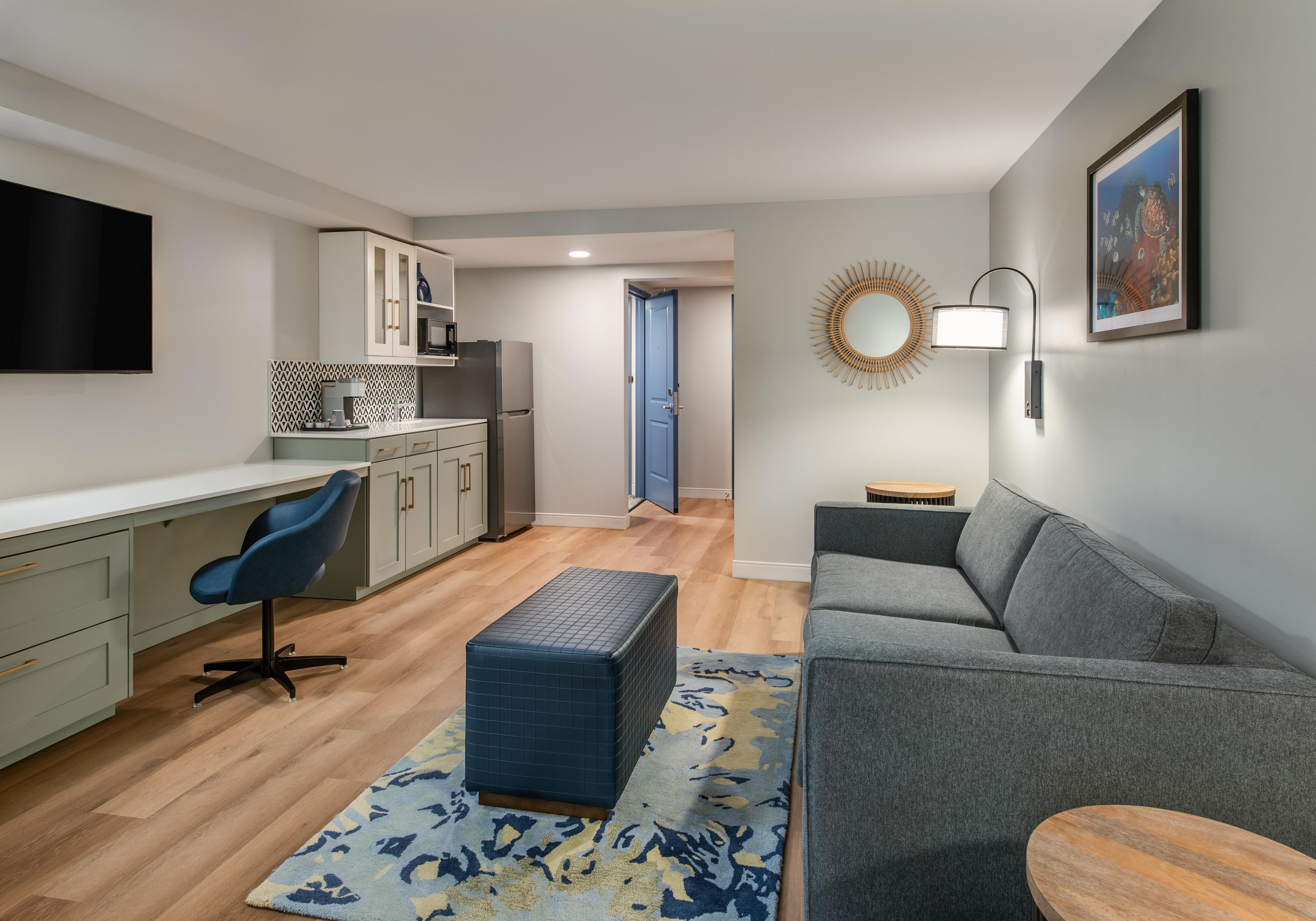 Ein modernes Wohnzimmer mit grauem Sofa, blauem Sessel, Holztisch, gemustertem Teppich und einer Küchenzeile mit Schränken und Fernseher.