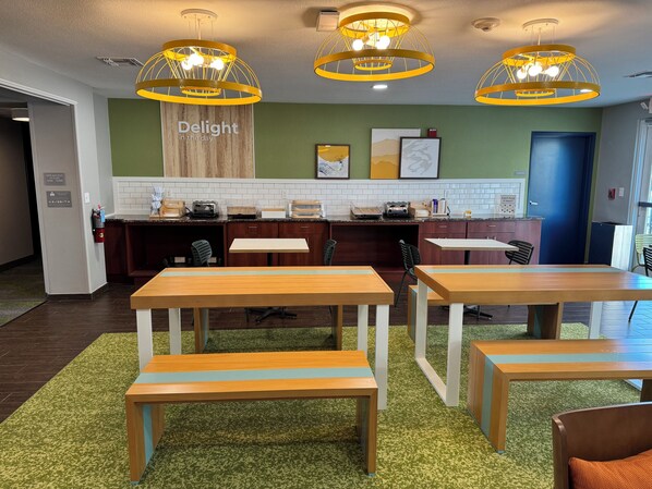 A modern dining area with wooden tables and benches, a kitchen counter with appliances, and a green wall with a 'Delight in the day' sign.