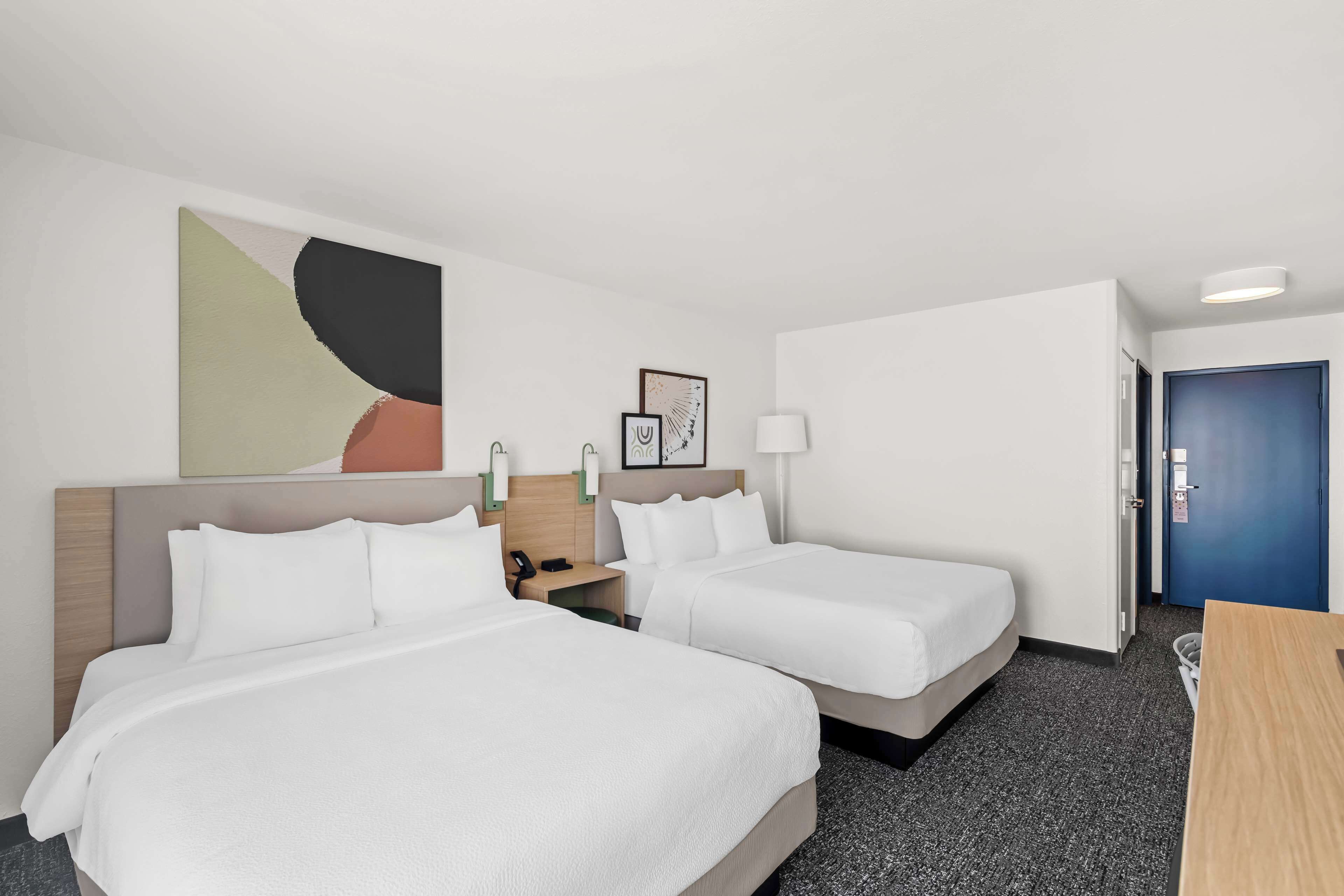 Quarto de hotel com duas camas, uma escrivaninha, uma luminária e uma porta.