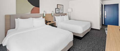 Quarto de hotel com duas camas, uma escrivaninha, uma luminária e uma porta.